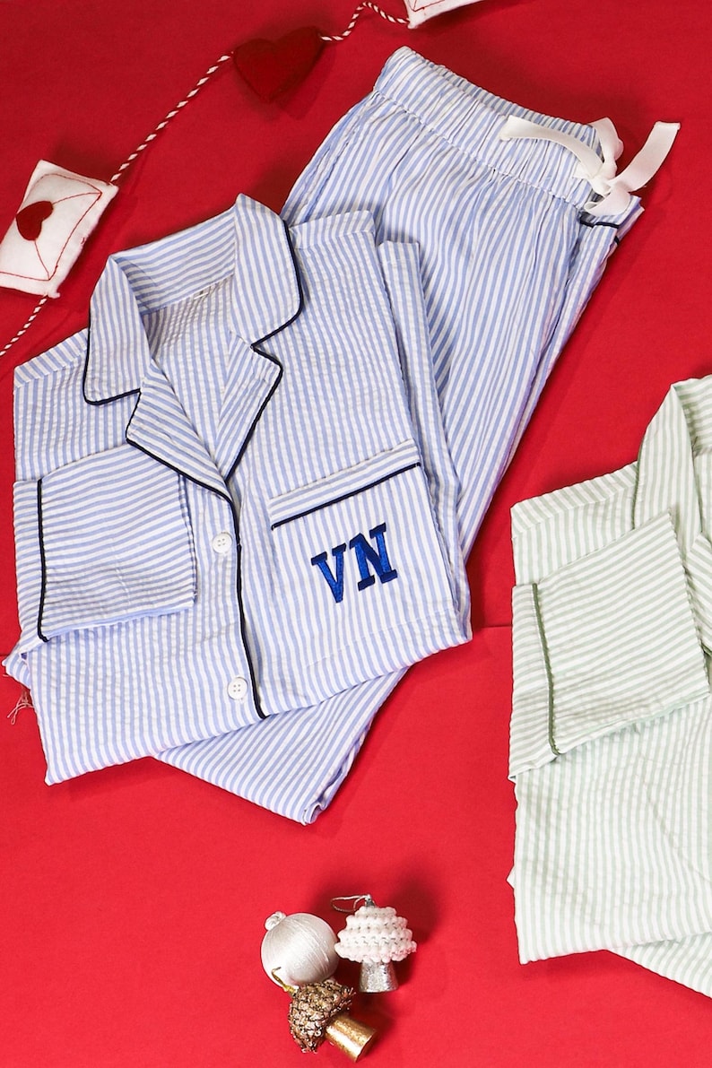 May include: A set of blue and white striped pajamas featuring a button-down shirt with a collar, long sleeves, and the letters VN embroidered in blue on the chest pocket. The matching pants have an elastic waistband with a white ribbon tie.