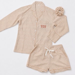 May include: A beige and white striped pajama set consisting of a long-sleeved button-down shirt with a collar and a chest pocket embroidered with the name TED in red, matching shorts with a white drawstring, and a coordinating fabric hair scrunchie.