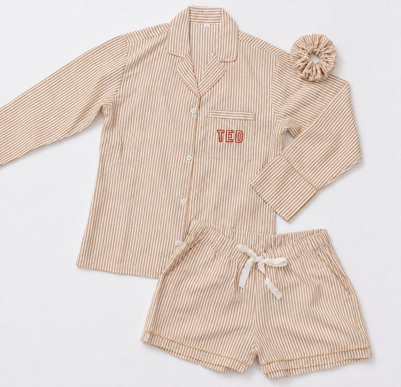 May include: A beige and white striped pajama set consisting of a long-sleeved button-down shirt with a collar and a chest pocket embroidered with the name TED in red, matching shorts with a white drawstring, and a coordinating fabric hair scrunchie.
