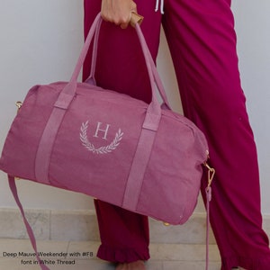 May include: A deep mauve weekender bag with a gold zipper and a white embroidered letter "H" with a laurel wreath design. The bag has a long strap and two top handles.