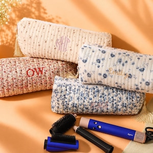May include: Four rectangular, quilted fabric pouches with floral patterns and tassel zipper pulls. Three pouches feature embroidered monograms or names, including KHP, OW, and Emily. A blue and copper hair styling tool with attachments lies below.