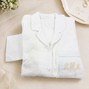 May include: A white long-sleeved button-down pajama top featuring a subtle vertical striped texture and a notched collar. The garment includes a single chest pocket embroidered with gold-colored cursive monogram letters and matching white buttons.