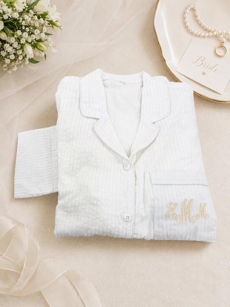 May include: A white long-sleeved button-down pajama top featuring a subtle vertical striped texture and a notched collar. The garment includes a single chest pocket embroidered with gold-colored cursive monogram letters and matching white buttons.