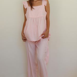 Op de afbeelding: Een lichtroze pyjamaset. De set bevat een top met ruchesbandjes en een bijpassende broek met wijde pijpen. De broek heeft een gefranjerde zoom. Het model draagt lichtroze slippers.