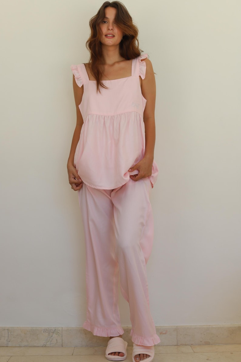 Op de afbeelding: Een lichtroze pyjamaset. De set bevat een top met ruchesbandjes en een bijpassende broek met wijde pijpen. De broek heeft een gefranjerde zoom. Het model draagt lichtroze slippers.