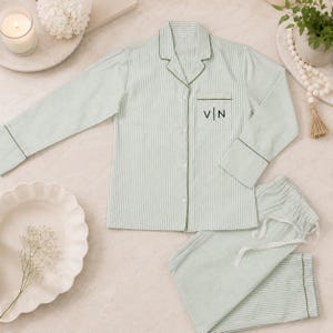 May include: A light green and white pinstriped pajama set consisting of a long-sleeved button-down shirt with a notched collar and a chest pocket featuring the embroidered black letters V | N, paired with matching drawstring pajama pants.