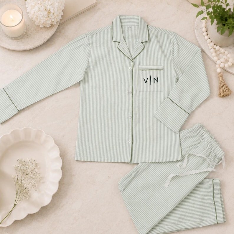 May include: A light green and white pinstriped pajama set consisting of a long-sleeved button-down shirt with a notched collar and a chest pocket featuring the embroidered black letters V | N, paired with matching drawstring pajama pants.