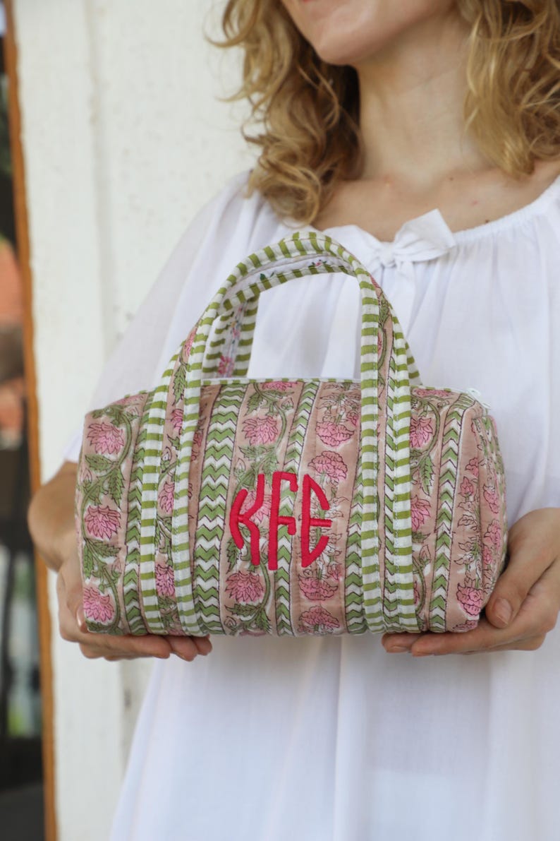 May include: A small, quilted handbag with a floral pattern in pink and green. The bag features a red monogram and green and white striped accents. The handles are also green and white striped. The bag is held by a person wearing a white top.