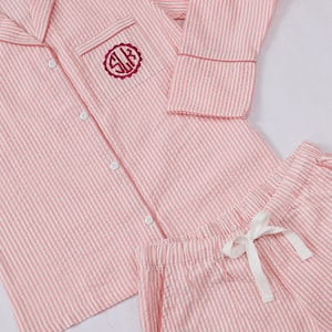 May include: A pink and white striped pajama set featuring a long-sleeved button-down shirt with a notched collar and a chest pocket. The pocket displays a circular dark pink monogram with the letters SLK. Matching shorts have a white drawstring.