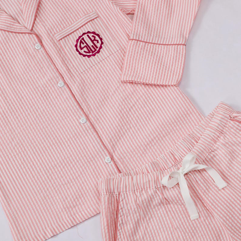 May include: A pink and white striped pajama set featuring a long-sleeved button-down shirt with a notched collar and a chest pocket. The pocket displays a circular dark pink monogram with the letters SLK. Matching shorts have a white drawstring.