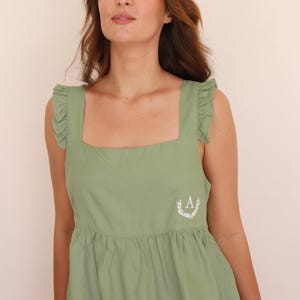 May include: Sage green dress with square neckline and ruffle details on the straps. Features a white embroidered "A" within a laurel wreath on the bodice. The dress has a gathered skirt and is made of a soft fabric.