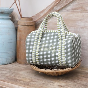 May include: A small, green and white gingham patterned bag with the name "Emily" embroidered on the front. The bag has a matching handle and is sitting in a small woven basket. Decorative vases are in the background.