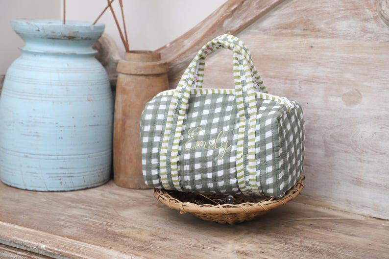 May include: A small, green and white gingham patterned bag with the name "Emily" embroidered on the front. The bag has a matching handle and is sitting in a small woven basket. Decorative vases are in the background.