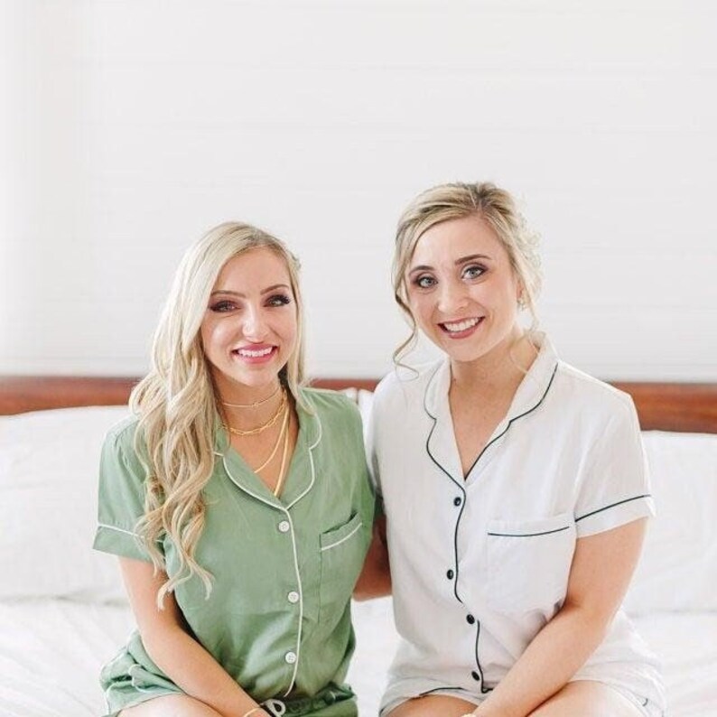 Op de afbeelding: Twee vrouwen in bijpassende pyjama's, een in groen en een in wit, met zwarte bies. Ze zitten op een bed en glimlachen naar de camera.