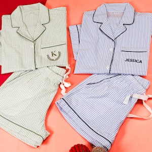 May include: Two sets of short-sleeved button-down pajamas with shorts. One set is light green with white stripes and a letter K embroidered on the pocket. The other set is light blue with white stripes and the name JESSICA embroidered on the pocket.