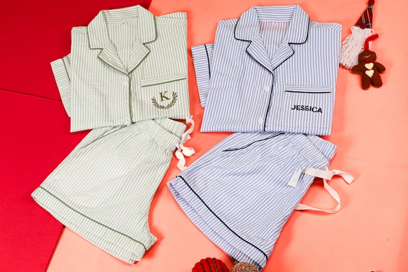 May include: Two sets of short-sleeved button-down pajamas with shorts. One set is light green with white stripes and a letter K embroidered on the pocket. The other set is light blue with white stripes and the name JESSICA embroidered on the pocket.