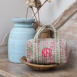 May include: A small, pink and green floral print duffel bag with a monogram in bright pink. The bag has a striped handle and is sitting in a woven basket. A light blue vase and dried flowers are in the background.