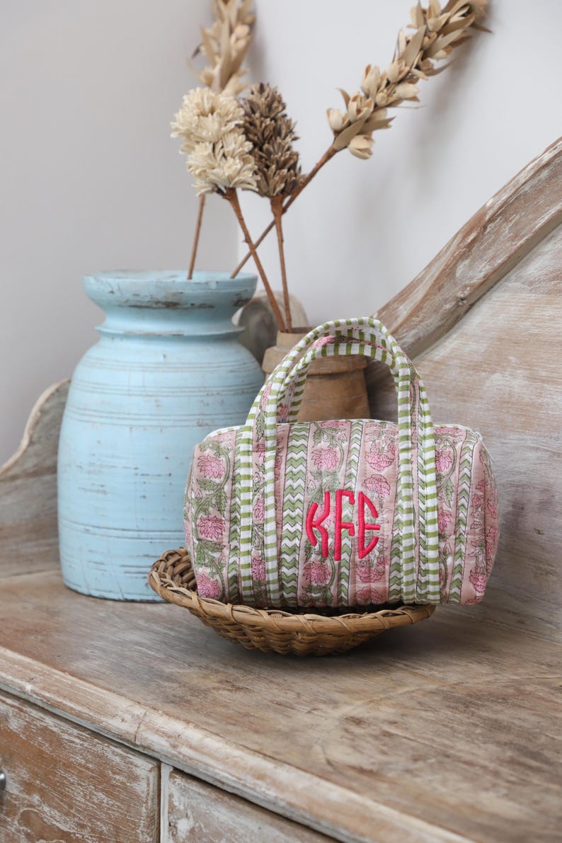 May include: A small, pink and green floral print duffel bag with a monogram in bright pink. The bag has a striped handle and is sitting in a woven basket. A light blue vase and dried flowers are in the background.
