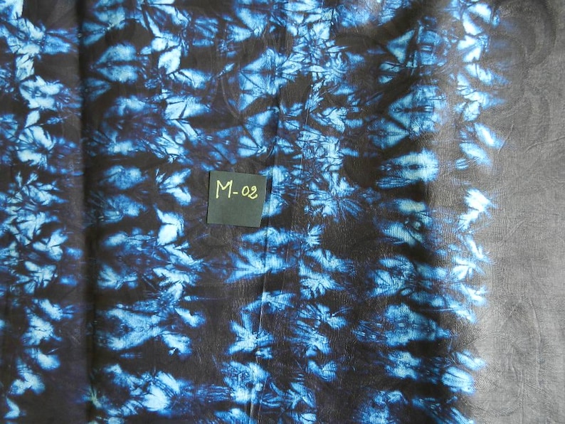 K&ouml;nnte beinhalten: Ein Stoff mit einem dunkelblauen und schwarzen Batikmuster. Der Stoff hat ein florales Design und den Text "M-02" auf einem kleinen Quadrat. Der Stoff wird wahrscheinlich f&uuml;r Kleidung oder Heimdekoration verwendet.
