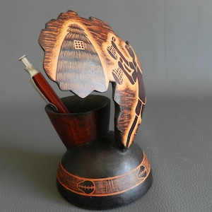 Puede incluir: Un portaplumas de madera con un diseño tallado de una cabaña y una persona. El portaplumas es marrón y negro con detalles en naranja. Un bolígrafo está en el portaplumas.