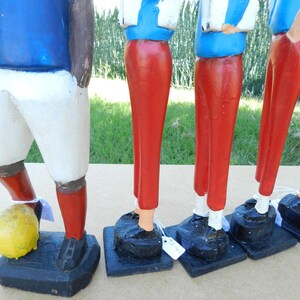 Puede incluir: Un conjunto de cinco figuras de madera pintadas a mano que representan jugadores de f&uacute;tbol. Cada figura viste una camiseta azul, pantalones rojos y zapatos negros sobre una base negra. Un jugador tiene un bal&oacute;n de f&uacute;tbol amarillo a sus pies.
