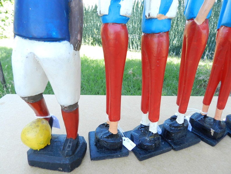 Puede incluir: Un conjunto de cinco figuras de madera pintadas a mano que representan jugadores de f&uacute;tbol. Cada figura viste una camiseta azul, pantalones rojos y zapatos negros sobre una base negra. Un jugador tiene un bal&oacute;n de f&uacute;tbol amarillo a sus pies.