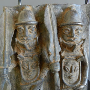 May include: A bronze plaque depicting two figures in a warrior pose, each holding a spear and wearing a helmet. The figures are carved in a detailed style with intricate facial features and clothing.