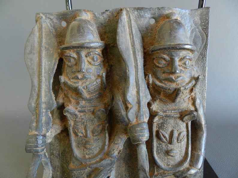 May include: A bronze plaque depicting two figures in a warrior pose, each holding a spear and wearing a helmet. The figures are carved in a detailed style with intricate facial features and clothing.