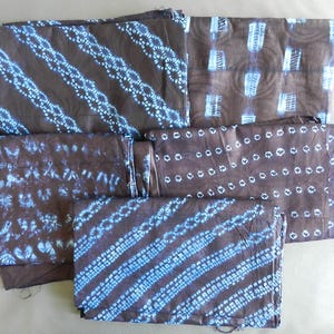 K&ouml;nnte beinhalten: Mehrere gefaltete Stoffst&uuml;cke in Braun- und Blaut&ouml;nen. Der Stoff weist verschiedene Muster auf, darunter diagonale Streifen, gepunktete Linien und abstrakte Formen. Die Designs sind in einem helleren Blau auf dunklerem braunen Hintergrund gehalten.