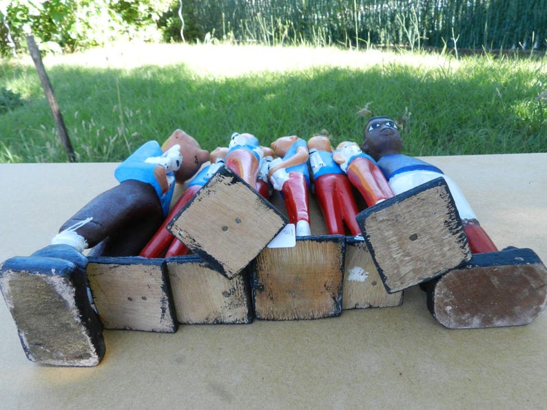 Puede incluir: Una colecci&oacute;n de seis figuras de madera pintadas a mano dispuestas en fila, cada una con un esquema de color &uacute;nico con camisas azules y pantalones rojos o blancos. Las figuras se ven desde un &aacute;ngulo bajo, destacando sus bases de madera cuadradas.