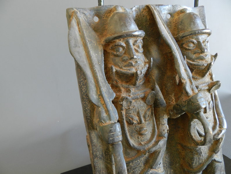May include: A bronze sculpture of two figures, each wearing a helmet and holding a spear. The figures are standing side-by-side and are facing forward. The sculpture is weathered and has a dark patina.