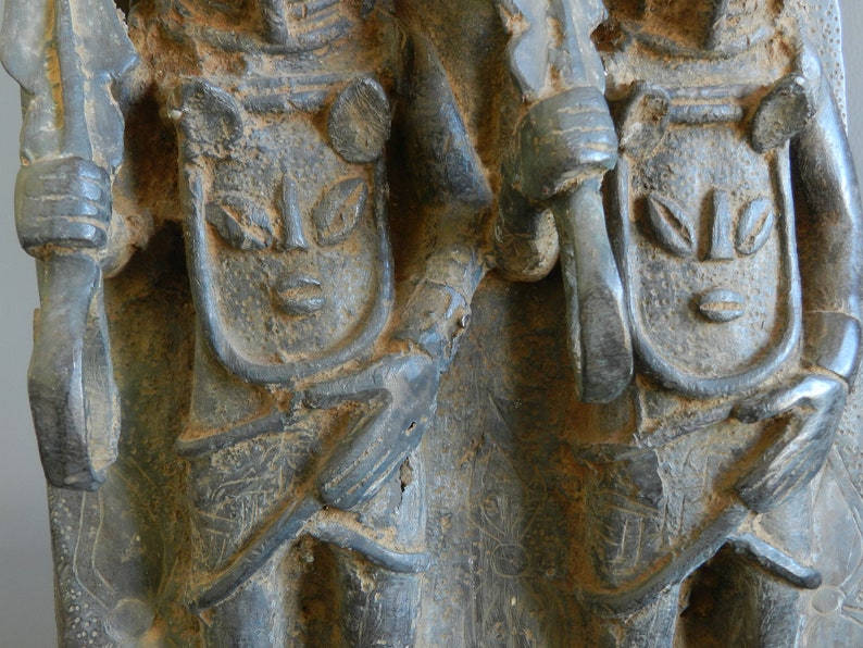 May include: Two carved stone figures with detailed faces and headdresses. The figures are holding spears and are standing side by side.