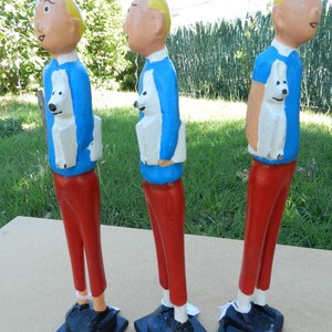 Puede incluir: Tres figuras de madera pintadas a mano de un personaje con pelo amarillo, camisa azul y pantalones rojos, cada una sosteniendo un perro blanco. Cada figura se apoya sobre una base negra con una etiqueta marcada como N&deg;001, N&deg;002 o N&deg;003.