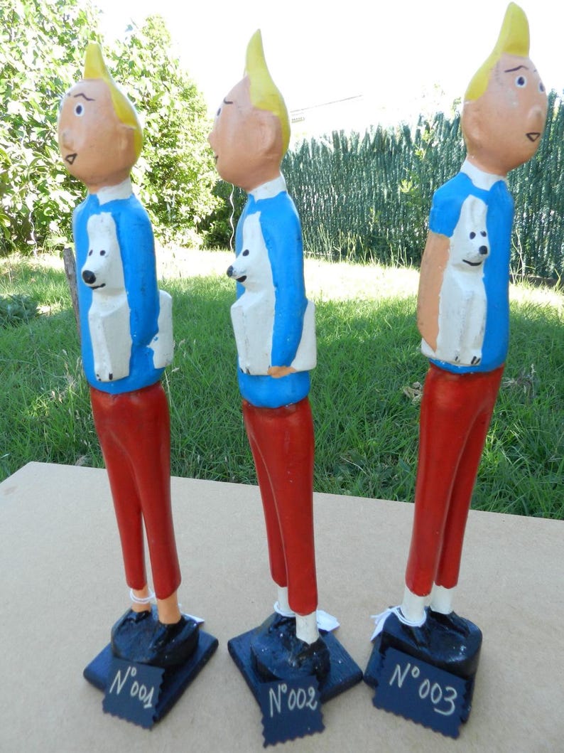 Puede incluir: Tres figuras de madera pintadas a mano de un personaje con pelo amarillo, camisa azul y pantalones rojos, cada una sosteniendo un perro blanco. Cada figura se apoya sobre una base negra con una etiqueta marcada como N&deg;001, N&deg;002 o N&deg;003.