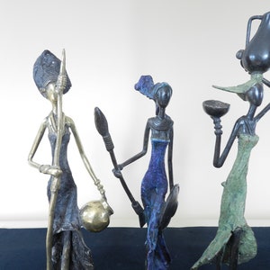Op de afbeelding: Vier bronzen sculpturen van vrouwen in traditionele Afrikaanse kleding. De sculpturen zijn gestileerd en abstract, met lange ledematen en overdreven kenmerken. De vrouwen houden verschillende voorwerpen vast, waaronder een kruik, een kom en een speer.