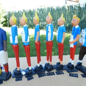 Puede incluir: Una colecci&oacute;n de ocho figuras pintadas a mano sobre bases negras. Una figura viste camisa azul y pantalones cortos blancos con un bal&oacute;n amarillo. Siete figuras muestran un personaje de pelo rubio, camisa azul, pantalones rojos y un perro blanco.
