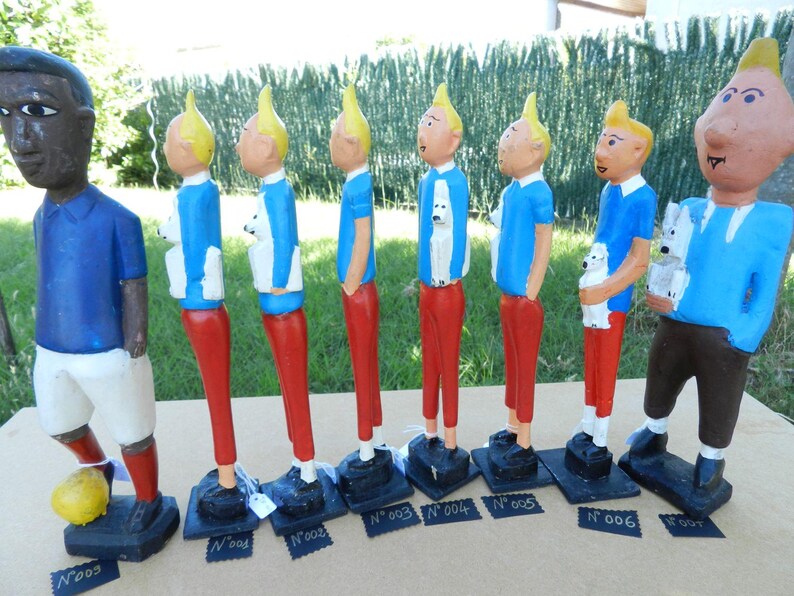 Puede incluir: Una colecci&oacute;n de ocho figuras pintadas a mano sobre bases negras. Una figura viste camisa azul y pantalones cortos blancos con un bal&oacute;n amarillo. Siete figuras muestran un personaje de pelo rubio, camisa azul, pantalones rojos y un perro blanco.