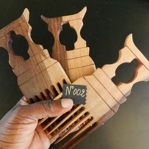 Puede incluir: Tres peines de madera con dientes largos y anchos, y un mango decorativo con un dise&ntilde;o recortado. La madera muestra vetas naturales de color marr&oacute;n. Una peque&ntilde;a etiqueta negra en el centro muestra el texto escrito a mano N&deg;002.