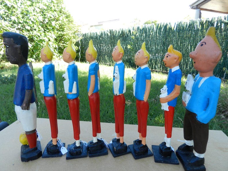 Puede incluir: Una colecci&oacute;n de ocho figuras de madera pintadas a mano dispuestas en fila. Siete figuras muestran un personaje con cabello amarillo, camisa azul y pantalones rojos, algunas sostienen un perro blanco. Una figura es de piel oscura y viste camisa azul.