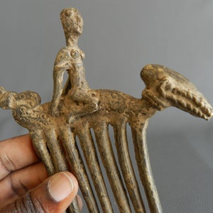 May include: A bronze comb with a figure of a person riding a stylized animal. The comb has a series of teeth and the figure is detailed with a raised design.