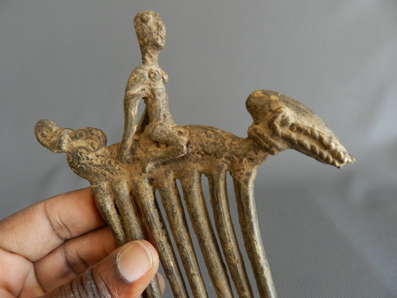 May include: A bronze comb with a figure of a person riding a stylized animal. The comb has a series of teeth and the figure is detailed with a raised design.