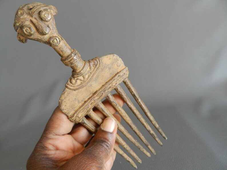 May include: A bronze comb with a detailed human-like head handle. The comb has seven teeth and is in a worn condition.