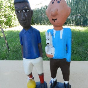 Puede incluir: Dos figuras de madera pintadas a mano sobre bases rectangulares. La figura de la izquierda viste camiseta azul, pantal&oacute;n corto blanco, calcetines rojos y un bal&oacute;n amarillo. La de la derecha tiene pelo rubio, camiseta azul y un perro blanco.