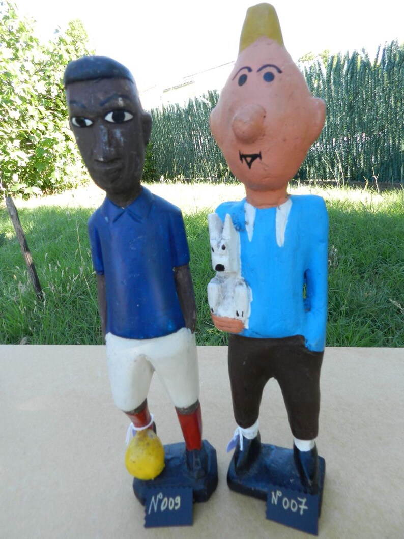 Puede incluir: Dos figuras de madera pintadas a mano sobre bases rectangulares. La figura de la izquierda viste camiseta azul, pantal&oacute;n corto blanco, calcetines rojos y un bal&oacute;n amarillo. La de la derecha tiene pelo rubio, camiseta azul y un perro blanco.