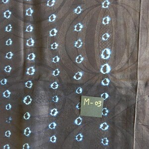 K&ouml;nnte beinhalten: Nahaufnahme eines dunkelbraunen Stoffes mit einem sich wiederholenden Muster aus wei&szlig;en und hellblauen Kreisformen. Der Stoff hat eine strukturierte Optik mit dezenten kreisf&ouml;rmigen Designs. Ein kleines schwarzes Quadrat mit dem Text "M-03" ist sichtbar.