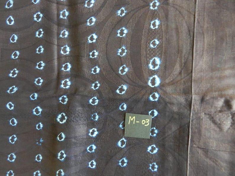 K&ouml;nnte beinhalten: Nahaufnahme eines dunkelbraunen Stoffes mit einem sich wiederholenden Muster aus wei&szlig;en und hellblauen Kreisformen. Der Stoff hat eine strukturierte Optik mit dezenten kreisf&ouml;rmigen Designs. Ein kleines schwarzes Quadrat mit dem Text "M-03" ist sichtbar.