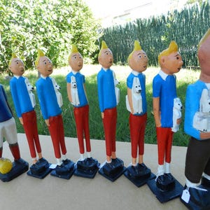 Puede incluir: Una colecci&oacute;n de ocho figuras de madera pintadas a mano dispuestas en fila. Siete figuras muestran a un personaje con cabello rubio, camisa azul y pantalones rojos, cada uno sosteniendo un perro blanco. Una figura viste camisa azul y pantalones cortos blancos con un bal&oacute;n amarillo.