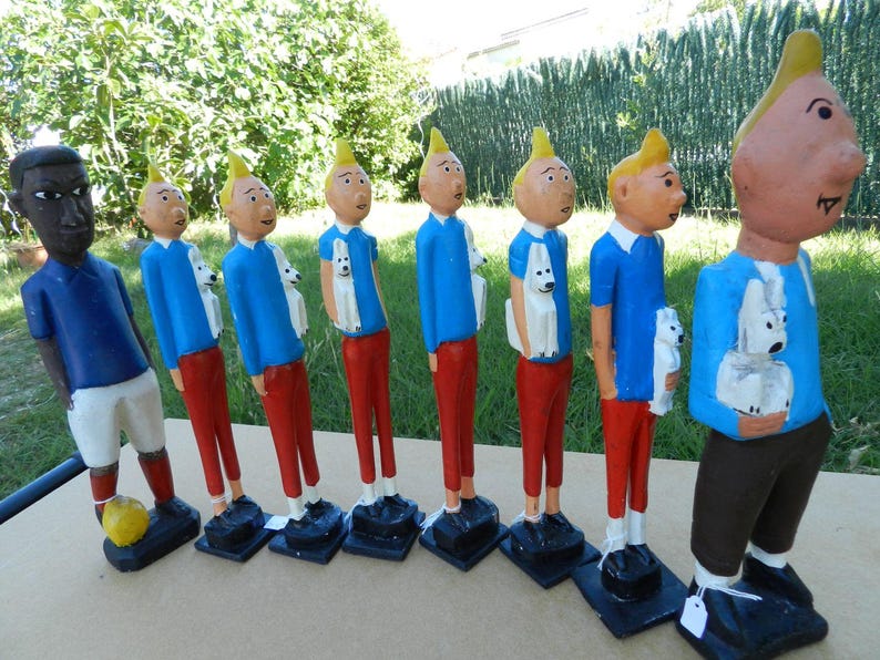 Puede incluir: Una colecci&oacute;n de ocho figuras de madera pintadas a mano dispuestas en fila. Siete figuras muestran a un personaje con cabello rubio, camisa azul y pantalones rojos, cada uno sosteniendo un perro blanco. Una figura viste camisa azul y pantalones cortos blancos con un bal&oacute;n amarillo.