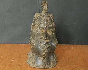 May include: A dark bronze sculpture of a human head with intricate details and a textured surface. The head is adorned with a crown and has a stylized face with large eyes and a prominent nose.