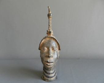 Peut inclure: Une sculpture en bronze d'une tête humaine avec une couronne haute et ornée. La tête est stylisée avec un visage plat et une bouche large et ouverte.
